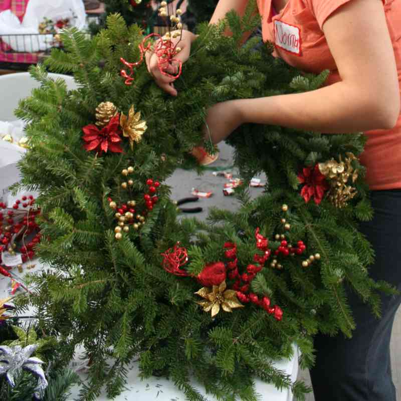 Person arranging a Christmas wreath with red and gold decorations on a table.