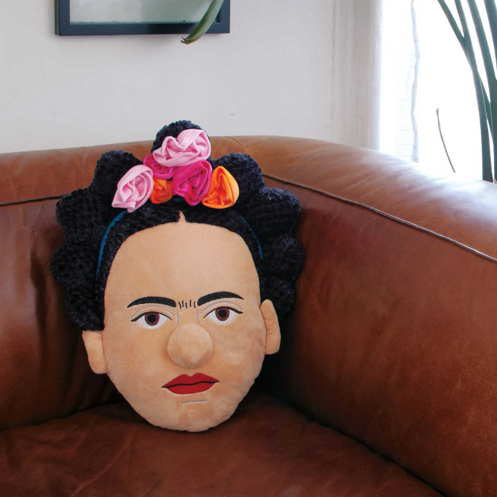 image of pillow in shape of Frida Kahlo's face sitting on the inside corner of a brown leather sofa