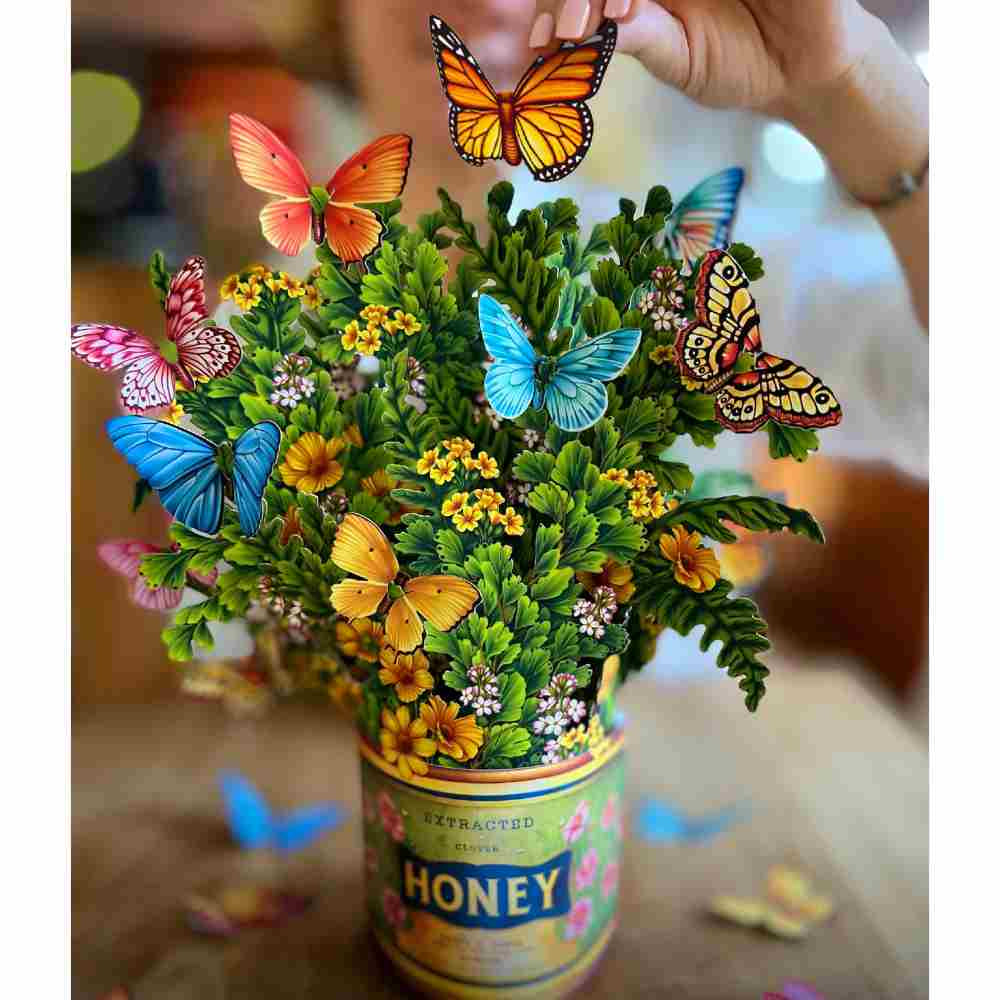 image of paper bouquet with multiple small yellow gold and light pink blossoms, along with several different colored butterflies adorning it.  A woman&#39;s hand is above the bouquet holding a monarch butterfly.  The vase is made to look like a vintage tin of honey