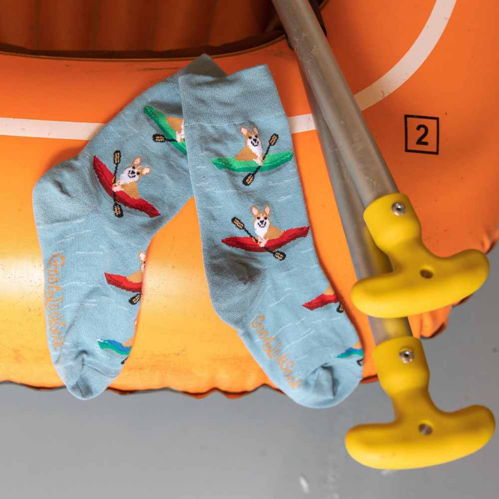 image of a pair of grey socks with drawings of corgi dogs in kayaks, hanging on the edge of a yellow inflatable raft next to the handles of oars