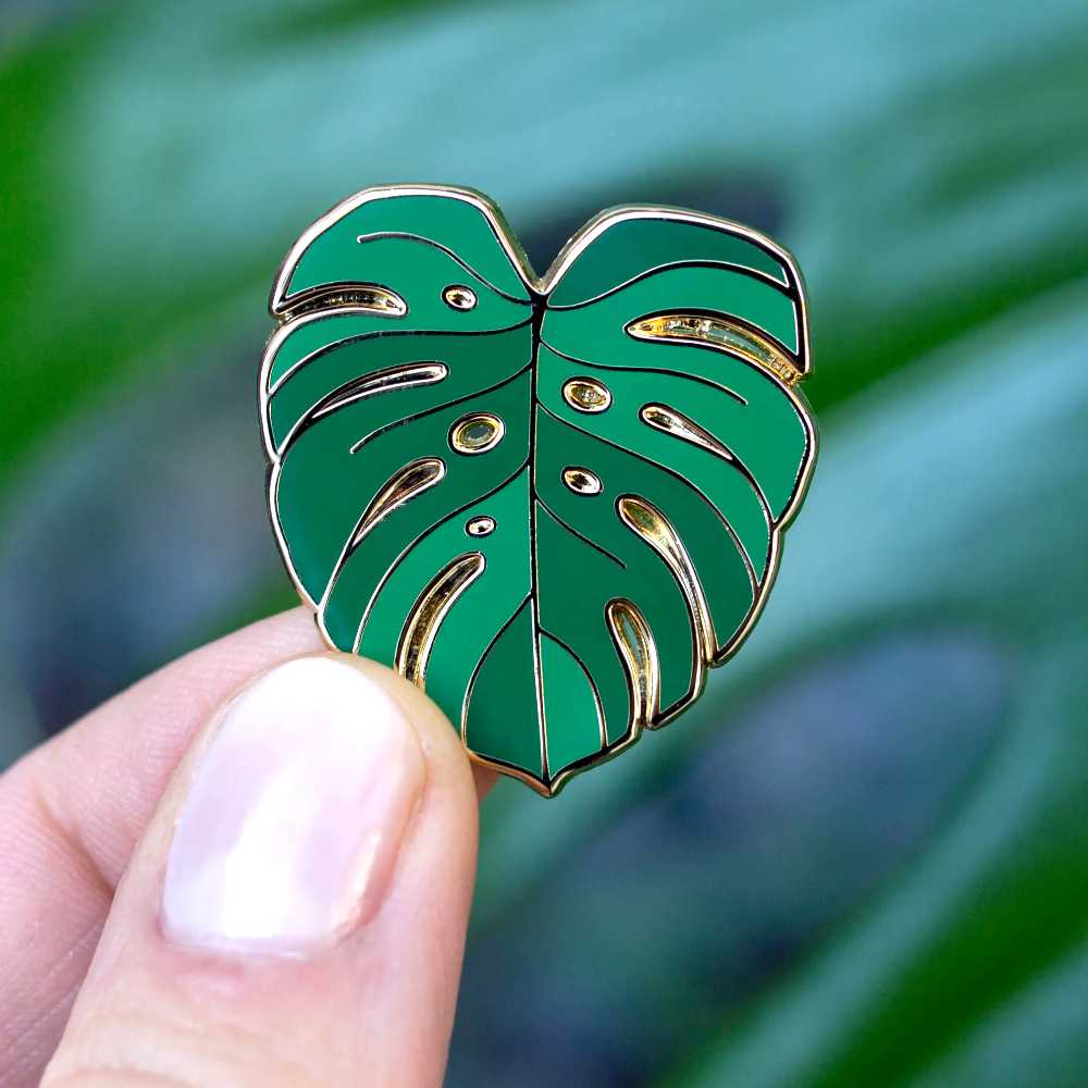 image of two fingers holding a small pin shaped like a monstera leaf, with two colors green and outlined in gold