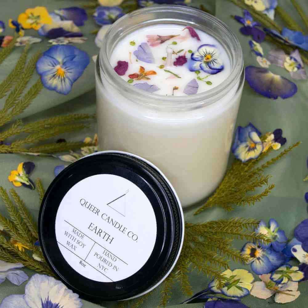 image of white candle in glass jar with small purple and red petals on top.  Black lid leaning again jar.  background surrounded by pressed multi color pansy blooms