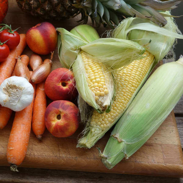 a cutting board with ears of corn, peaches, carrots, garlic, shallots and pineapple