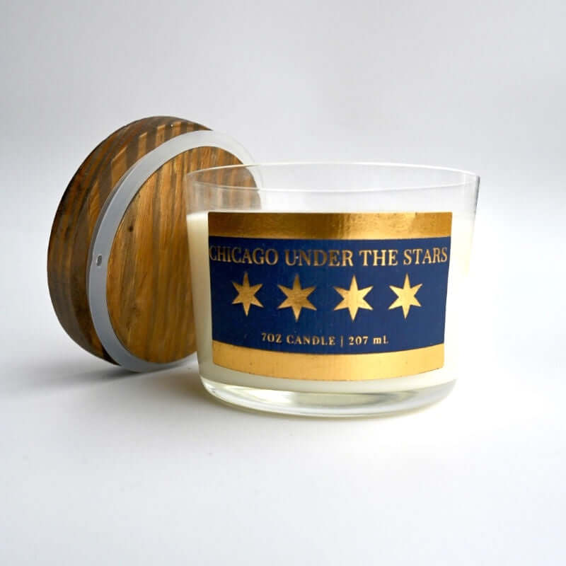 image of round clear glass jar candle with blue and gold labels featuring four 6 pointed stars.  Wood lid leaning against jar
