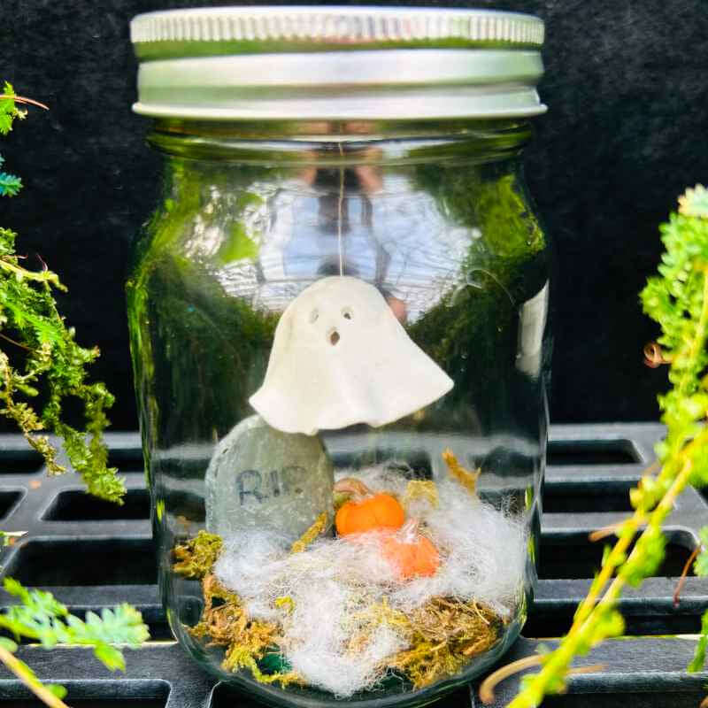 Mason jar with a small ghost sculpture, pumpkins, and headstone on a black background