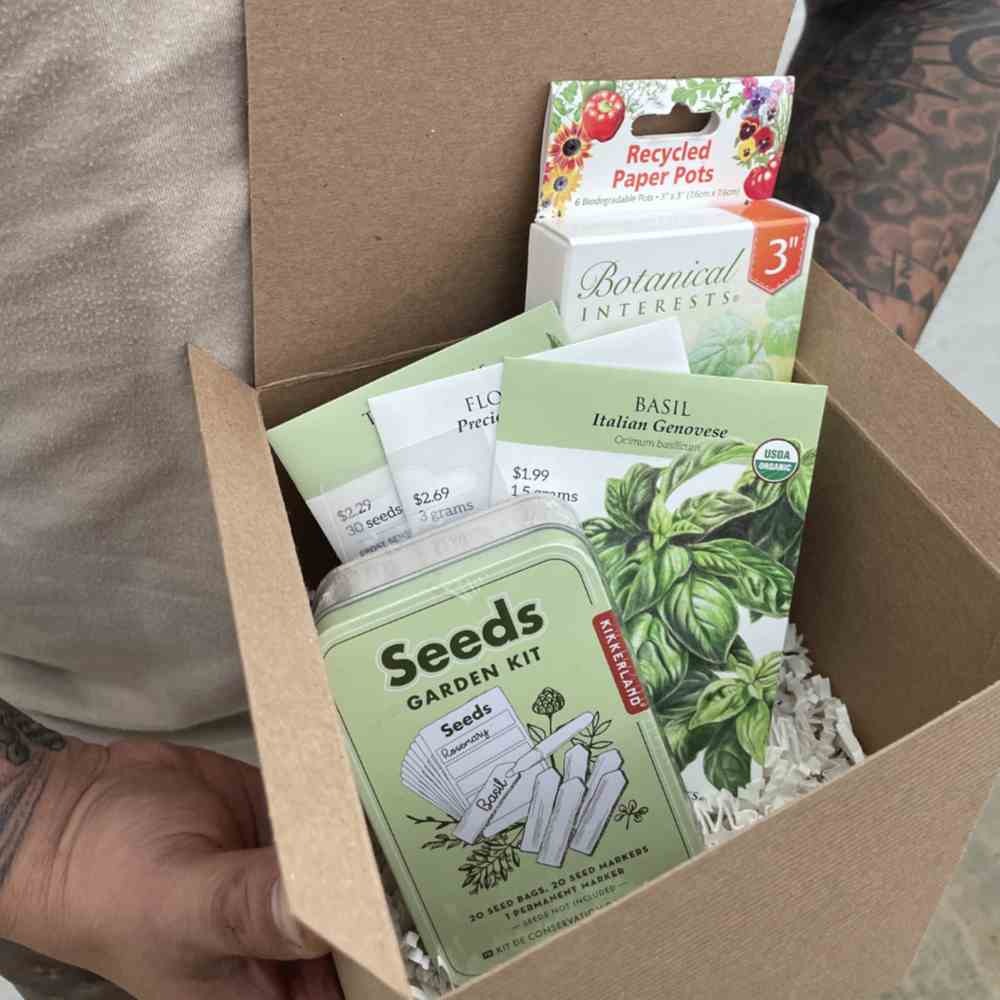 closeup image of person holding gifts box with Seeds Garden Kit tin, three seed packs and a cardboard package of recycled paper pots, all in a Kraft paper gift box with the top open