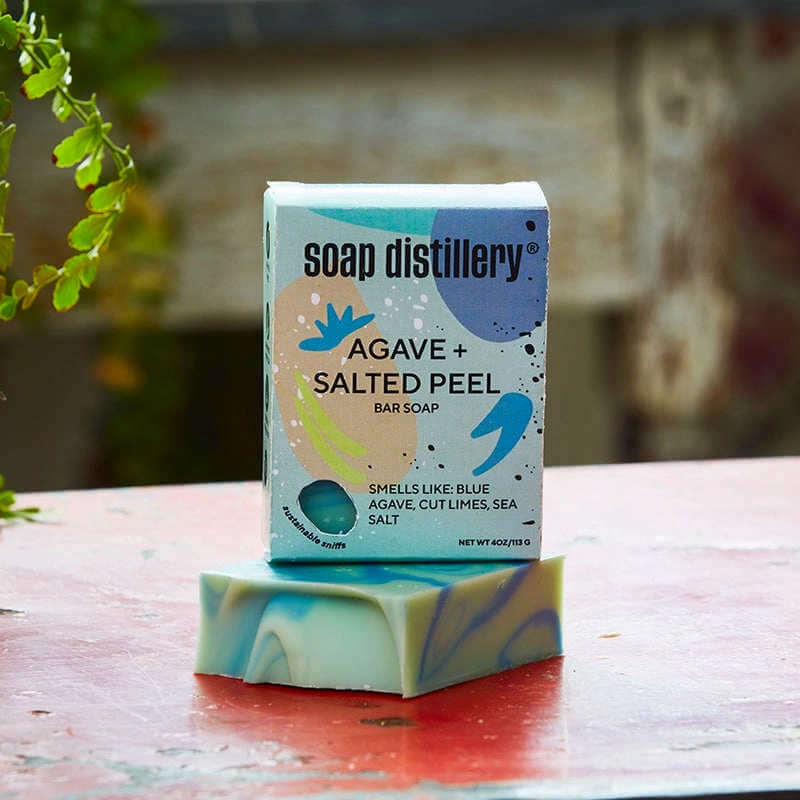 image of box in light blues and oranges with purple accents and the soap distillery logo sitting on top of a multicolor swirled bar of soap