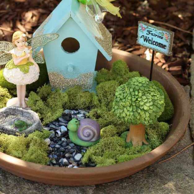 closeup image of a partial fairy garden with green moss, a birdhouse, a small fairy figure, a sign and other small figurines