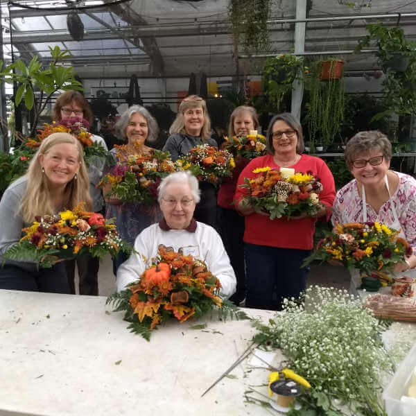 image of 8 women holding floral centerpieces in fall colors of orange and yellow, standing in front of a white table