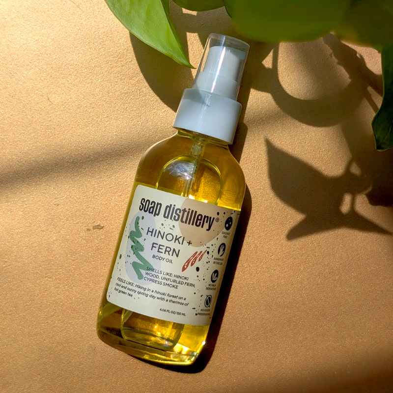 Bottle of Soap Distillery Hinoki Fern body oil on a beige background with plant leaves.