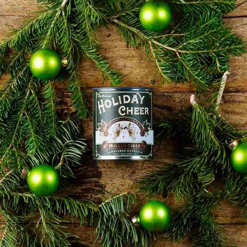 image of candle in round can with dark green and white label.  Surrounded by fresh evergreen boughs and round green ornaments