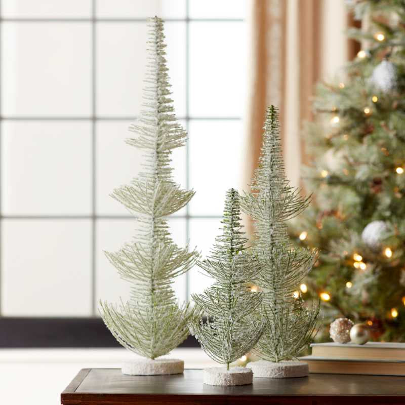 image of three different height bottlebrush style trees  covered in silvery glitter.  A Christmas tree is blurred in the background
