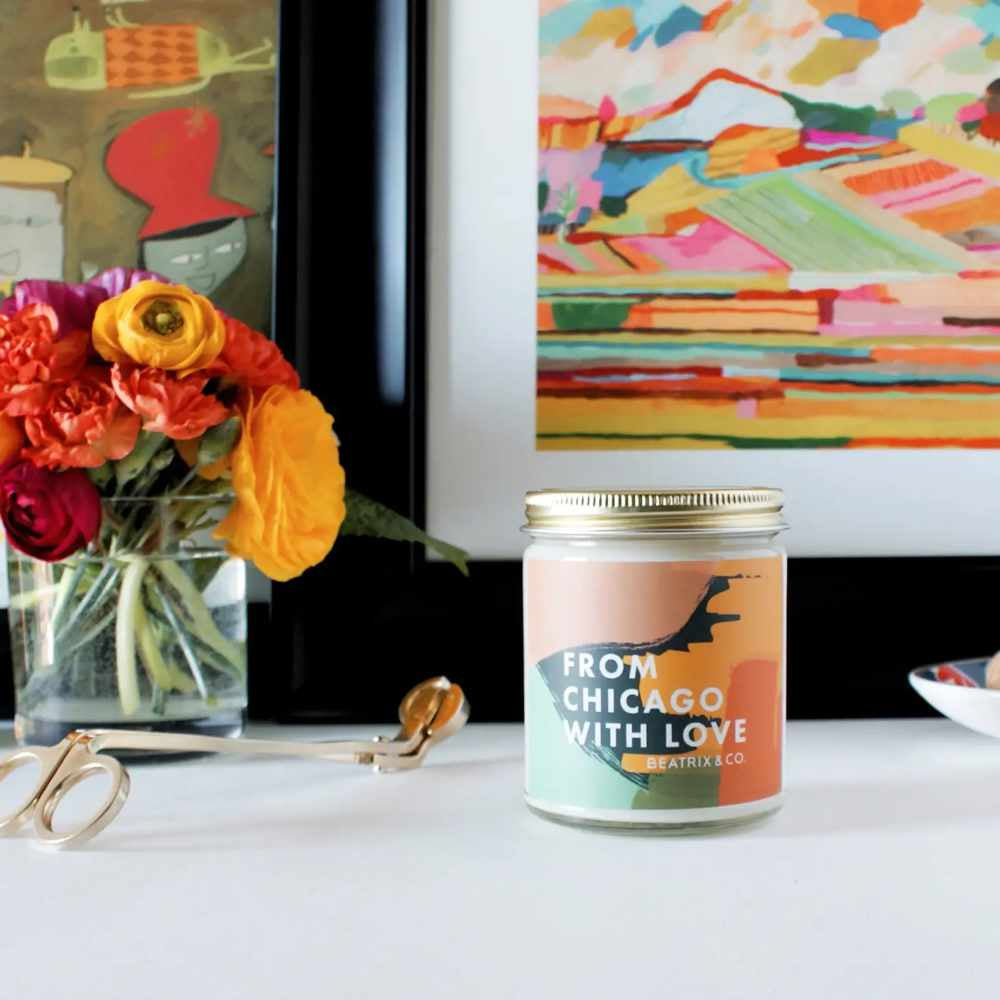 image of round jar candle with gold lid and abstract design on the label.  In background is a bouquet in yellow, red, pink and orange, as well as part of a painting in similar colors