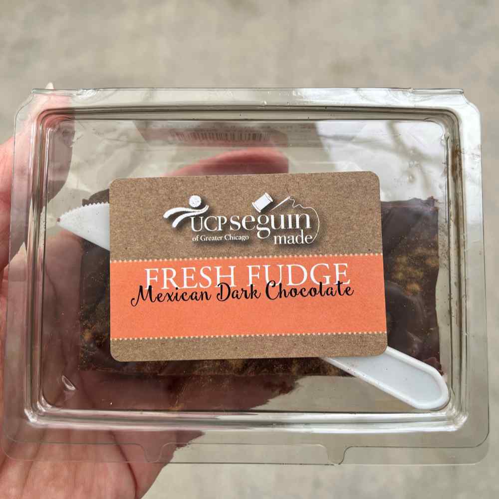 image of hand holding a clear plastic box with a piece of fudge in it.  Top of box has a label in tan and peach tones for Mexican Dark Chocolate fudge