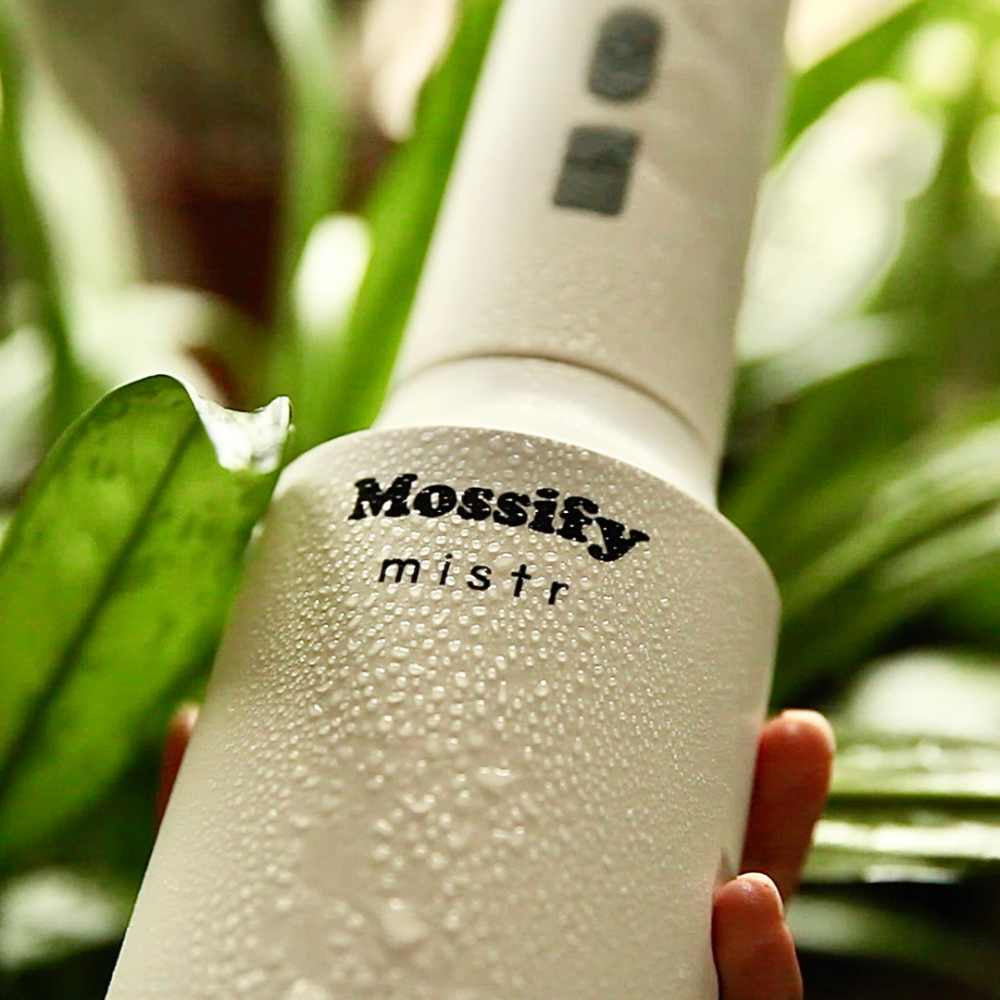 image of a hand holding a white cylindrical mister with a fine mist on it.  Logo on the front and green plants in the background