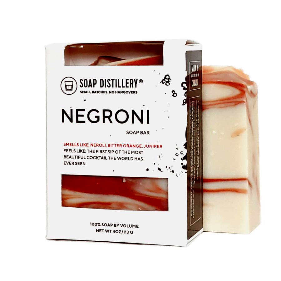 image of box of soap in white box with black text, and openings with view of soap bar inside.  Behind that is a bar of Negroni soap in cream and deep orange colors