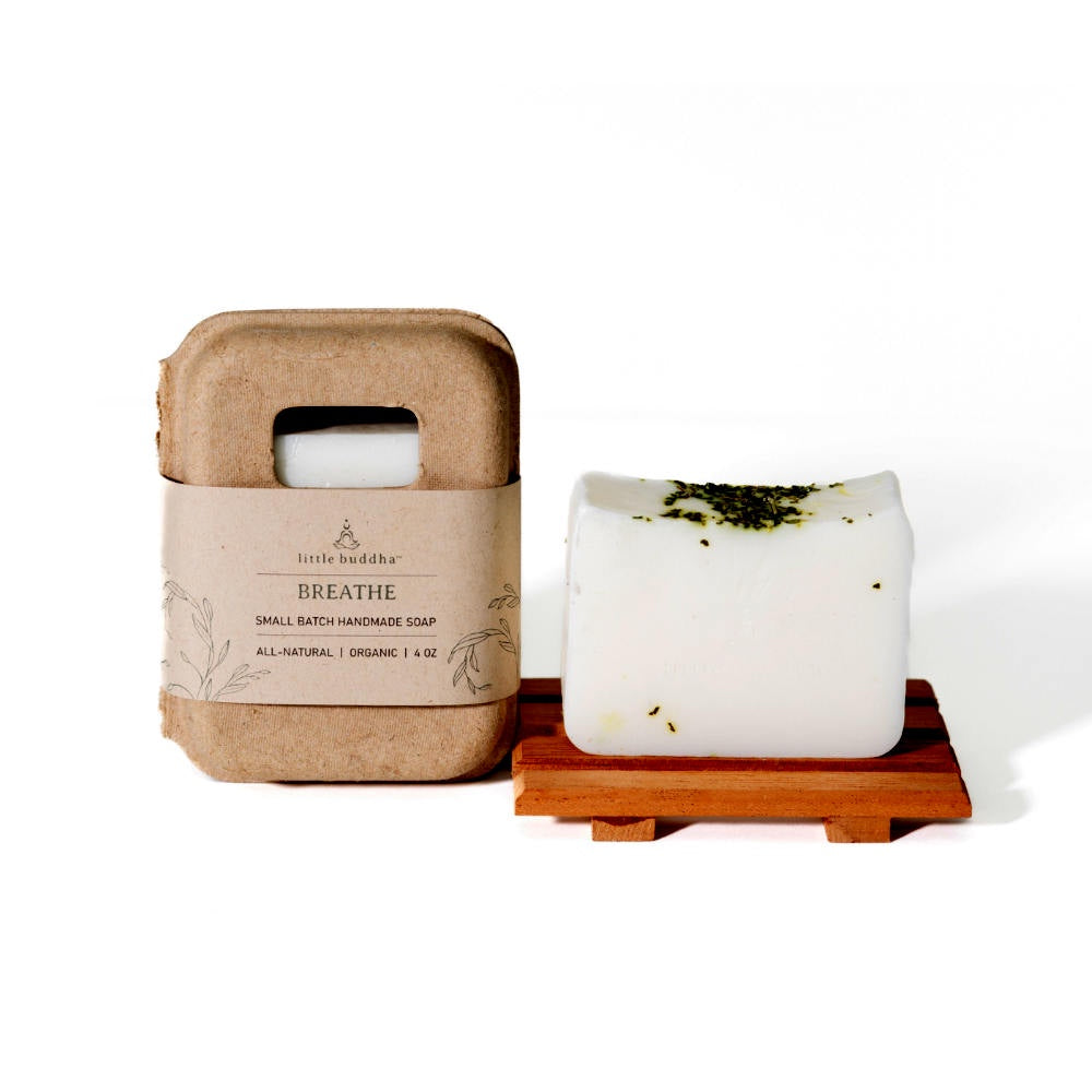 image of white soap sitting on a wooden soap dish.  To the left is the rounded cardboard box with kraft paper label that the soap comes in.