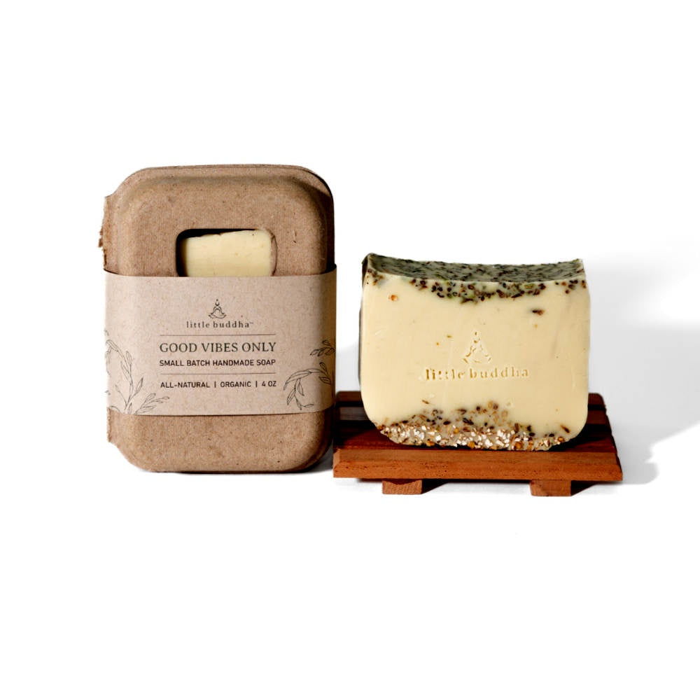 image of bar of soap in cream color with oatmeal accents on the top and bottom, sitting on a wood soap dish.  To the left is the rounded cardboard box with a kraft paper label that the soap comes in