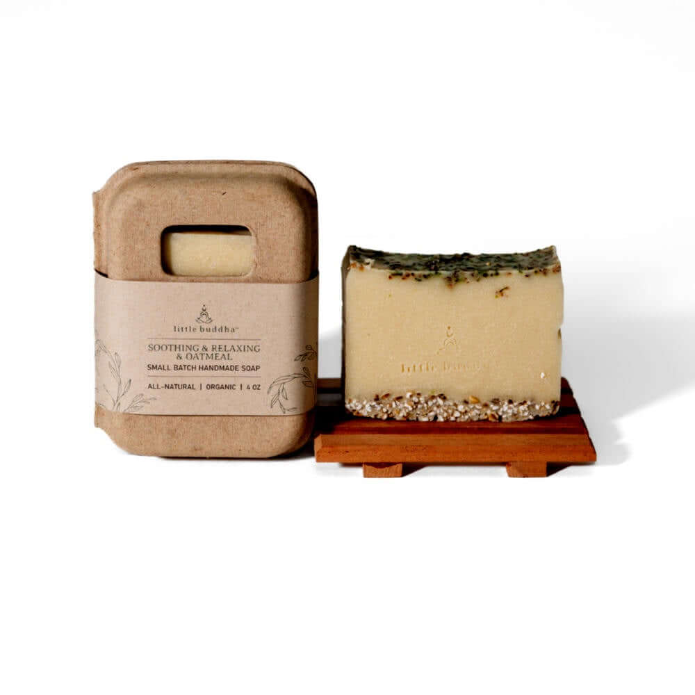 image of soap with oatmeal on the top and bottom edges, sitting on a wooden soap dish.  To the left is the rounded cardboard box with kraft paper label that the soap comes in.