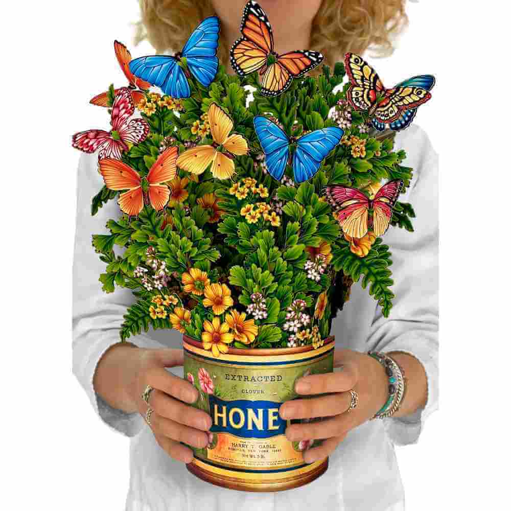 image of paper bouquet with multiple small yellow gold and light pink blossoms, along with several different colored butterflies adorning it.  A woman's hand is above the bouquet holding a monarch butterfly.  The vase is made to look like a vintage tin of honey