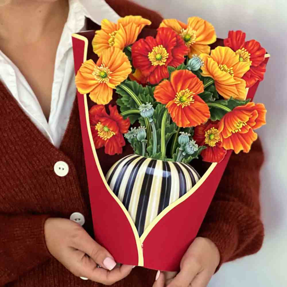 image of a woman&#39;s upper body and arm in a russet color sweater holding the opened bouquet, with a cream and black striped paper vase and large paper blossoms in yellow gold, light and dark orange.