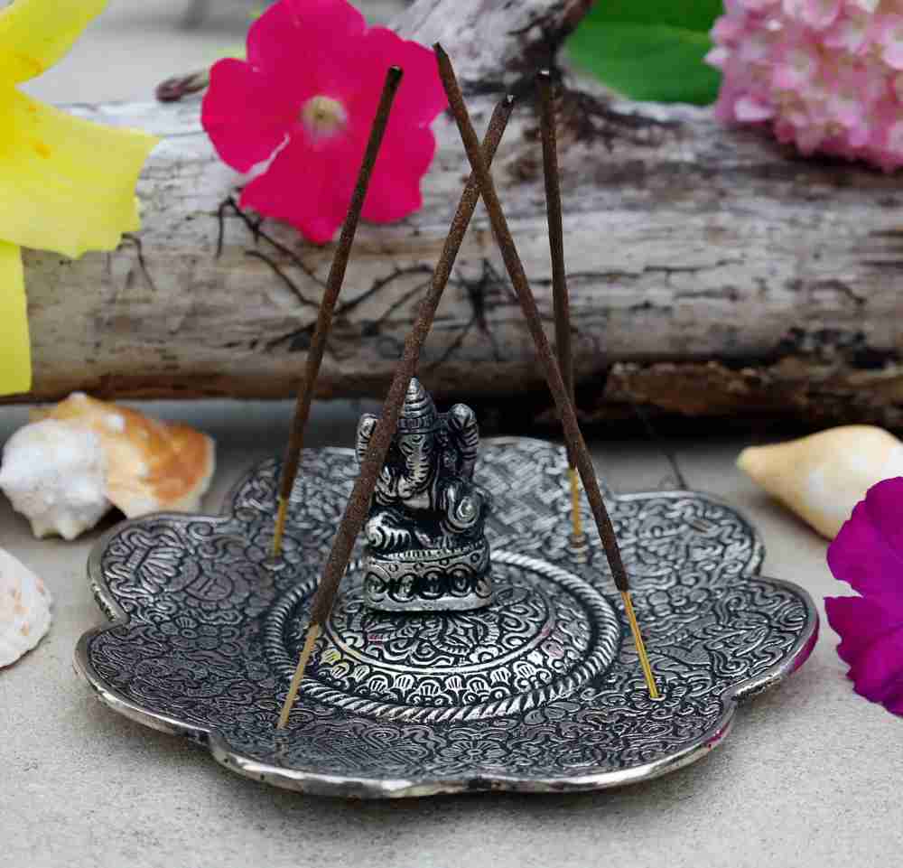 image of round scalloped silver platter with a small silver buddha at the center and four thin brown incense sticks sticking up from the disc.  flowers and log in background