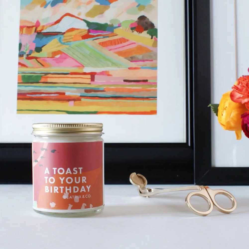 image of jar candle with red and orange abstract design on a counter next to a gold wick trimmer.  in the background is an abstract landscape planting in colors of orange, red, green, yellow and other colors