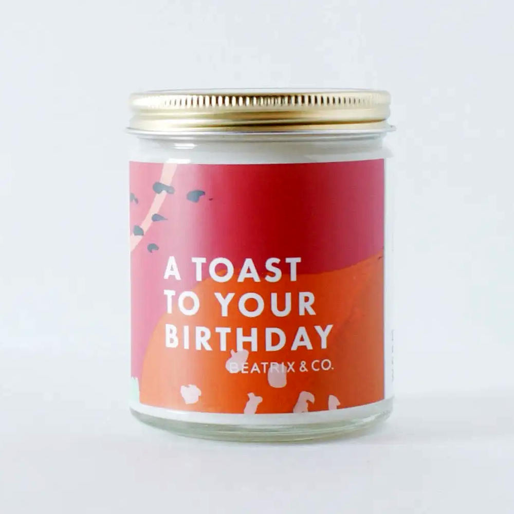 image of glass jar candle with gold lid.  Label is an abstract red and orange with type in white on top.
