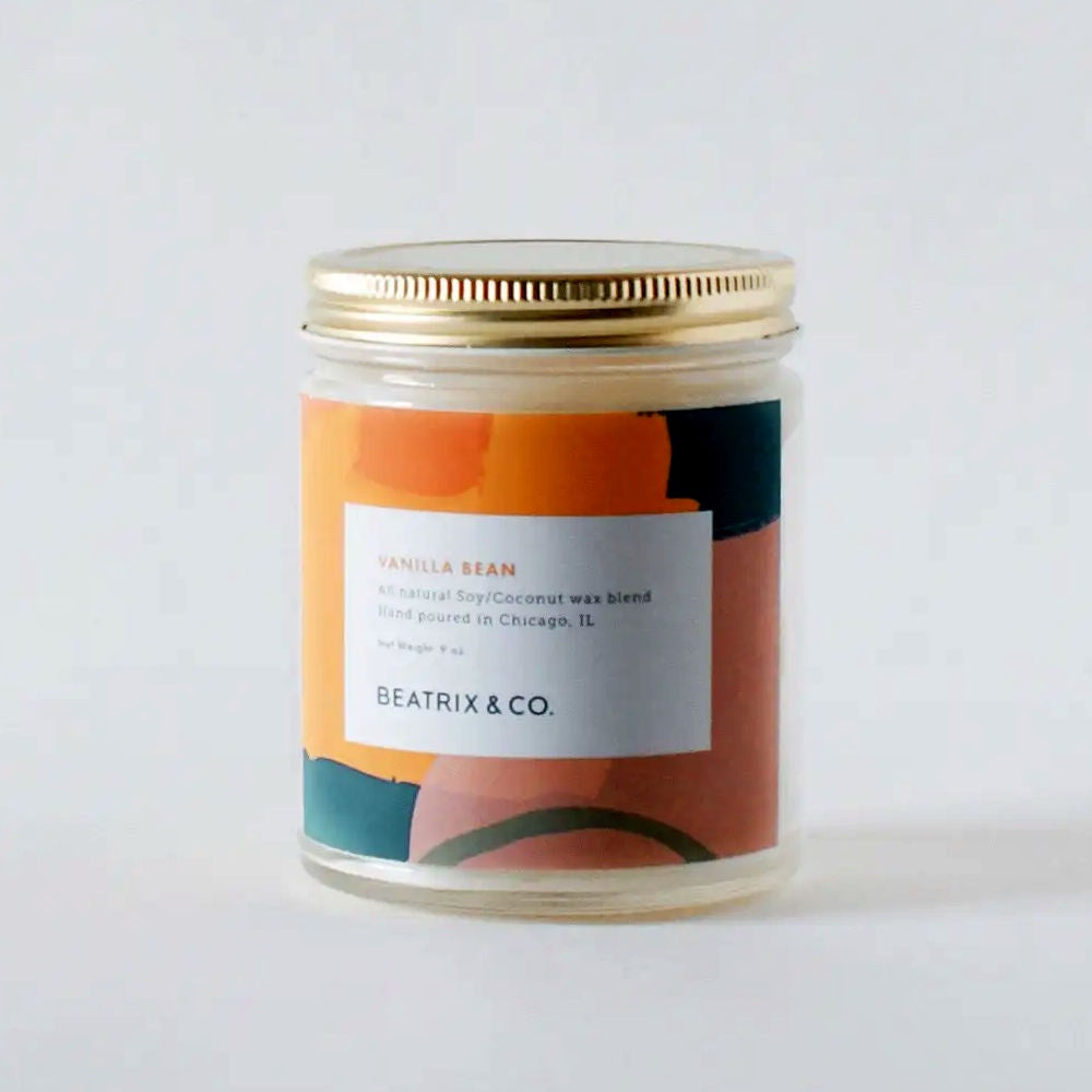 image of round glass jar candle with gold lid.  Label is abstract with colors of medium and deep orange, dark blue green, deep dusty pink and dark green.  In front of colors is a white rectangle with the candle description